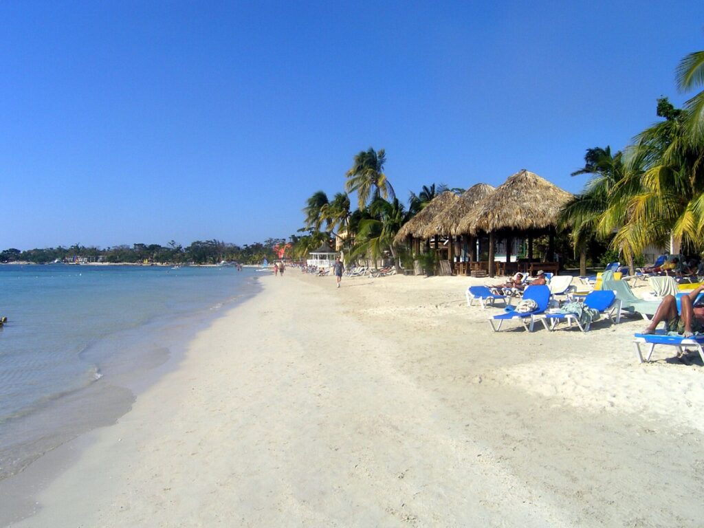 Long white-sand beach at Sandals Negril, Jamaica, with thatched-roof huts, blue loungers and palm trees beside calm turquoise water