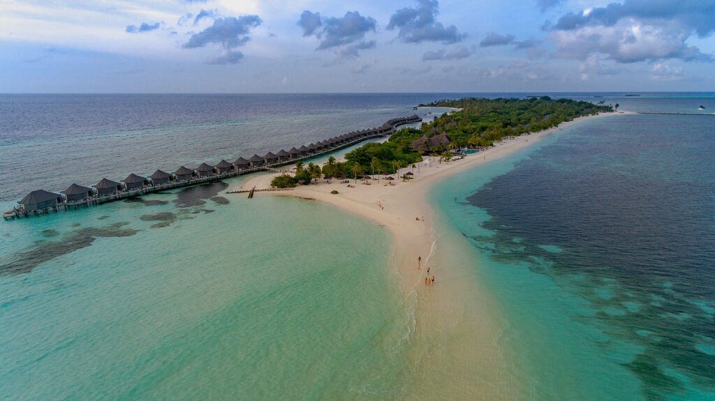 Aerial view of a long white sandbank curving into turquoise water beside a row of overwater villas in the Maldives
