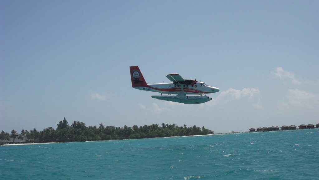 Trans Maldivian Airways seaplane coming in to land over a turquoise lagoon beside a palm-covered resort island in the Maldives