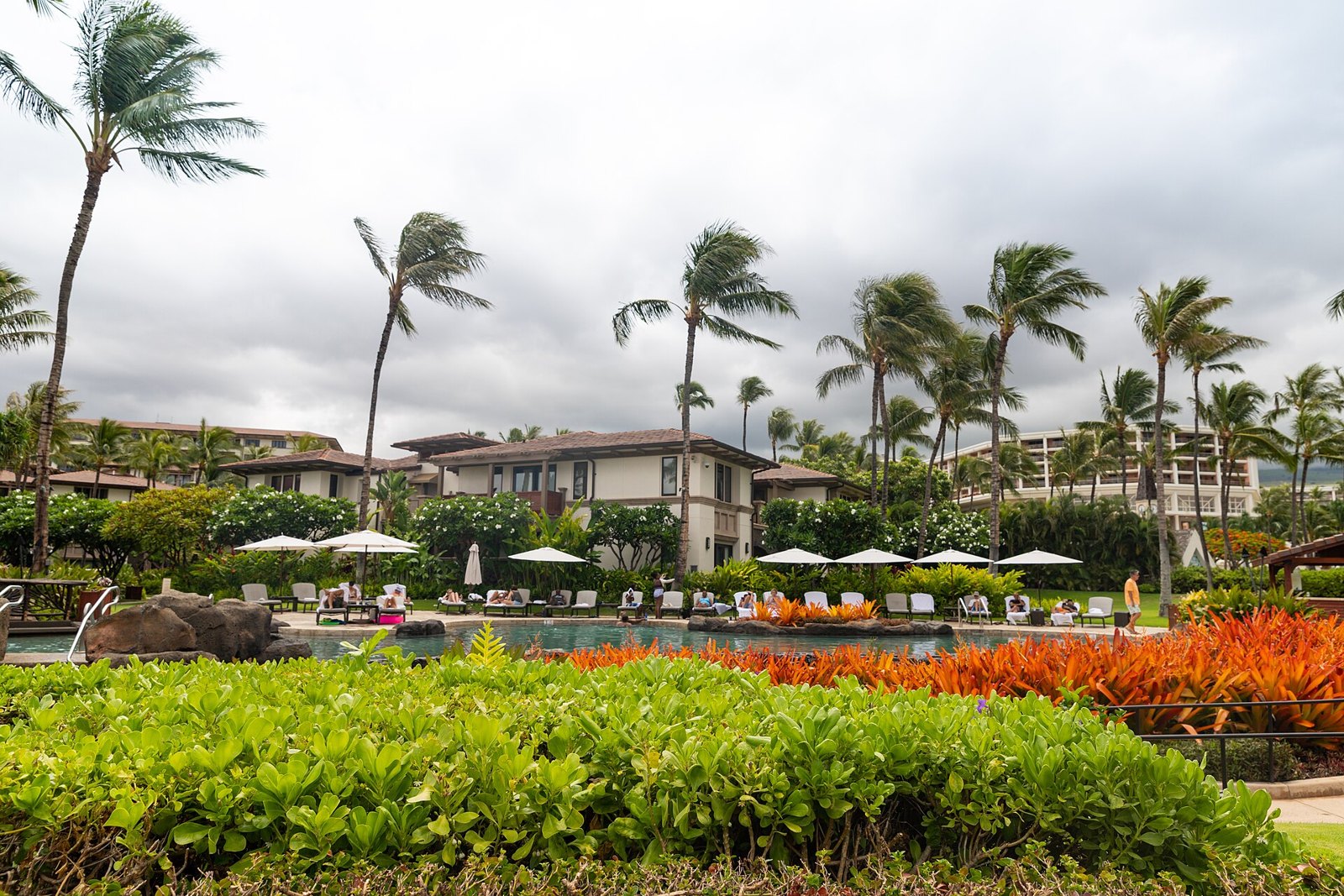 Resort pool deck with white umbrellas and palm trees under a cloudy sky in Wailea, Maui